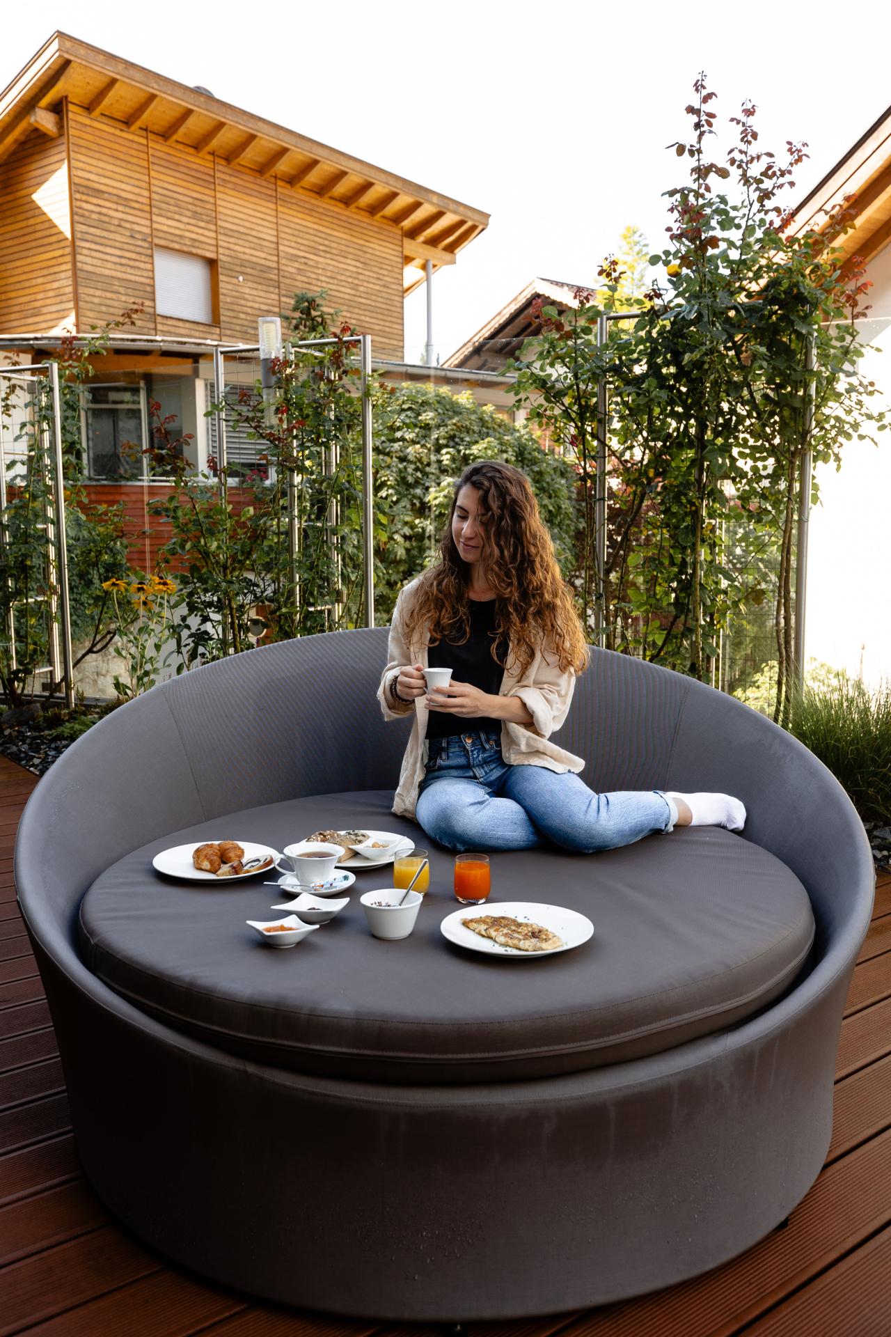 Frau, die in einem Garten auf einer runden Couch sitzt und frühstückt