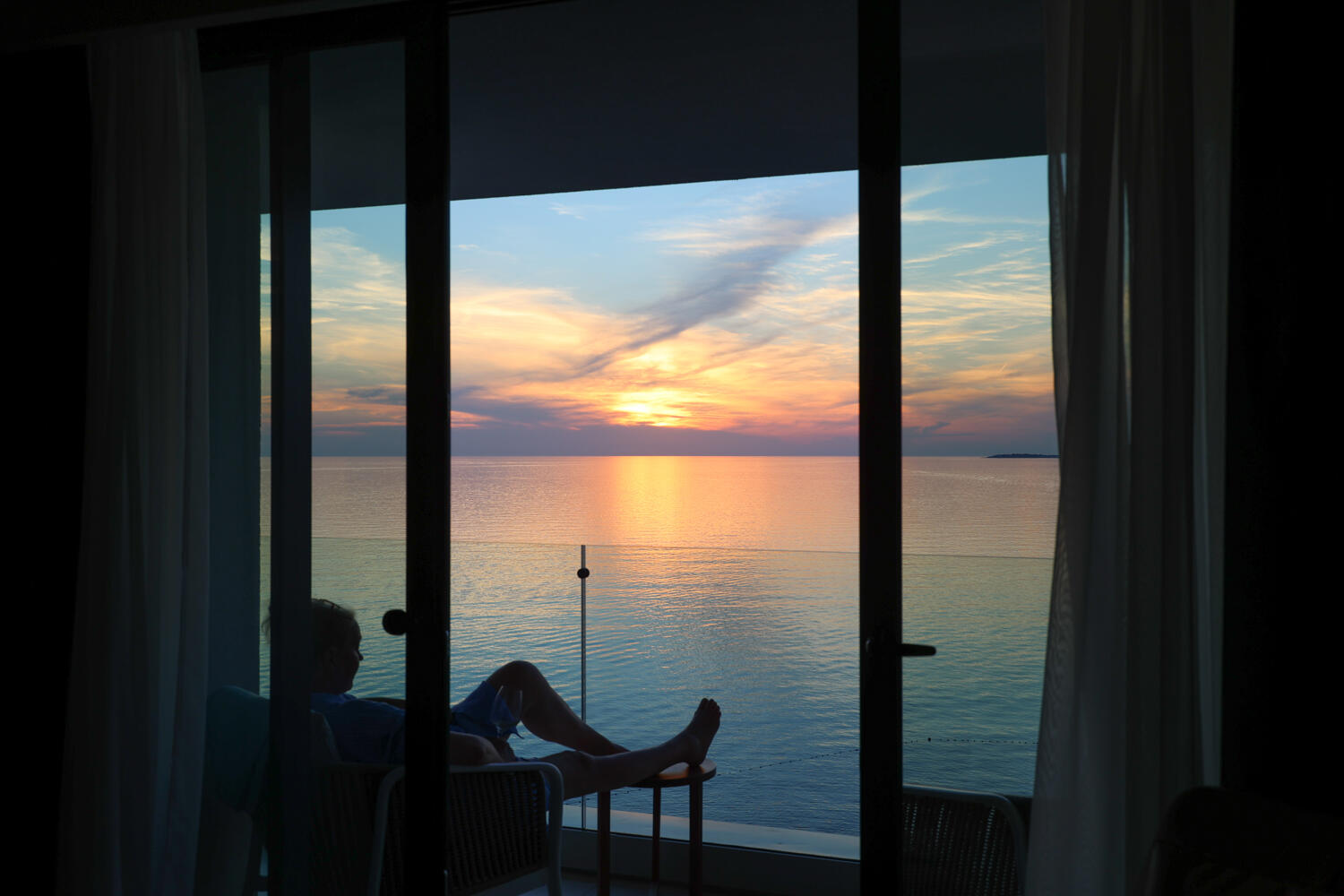 Frau, die in einem dunklen Zimmer mit Balkon sitzt und auf den Sonnenuntergang über dem Meer blickt.