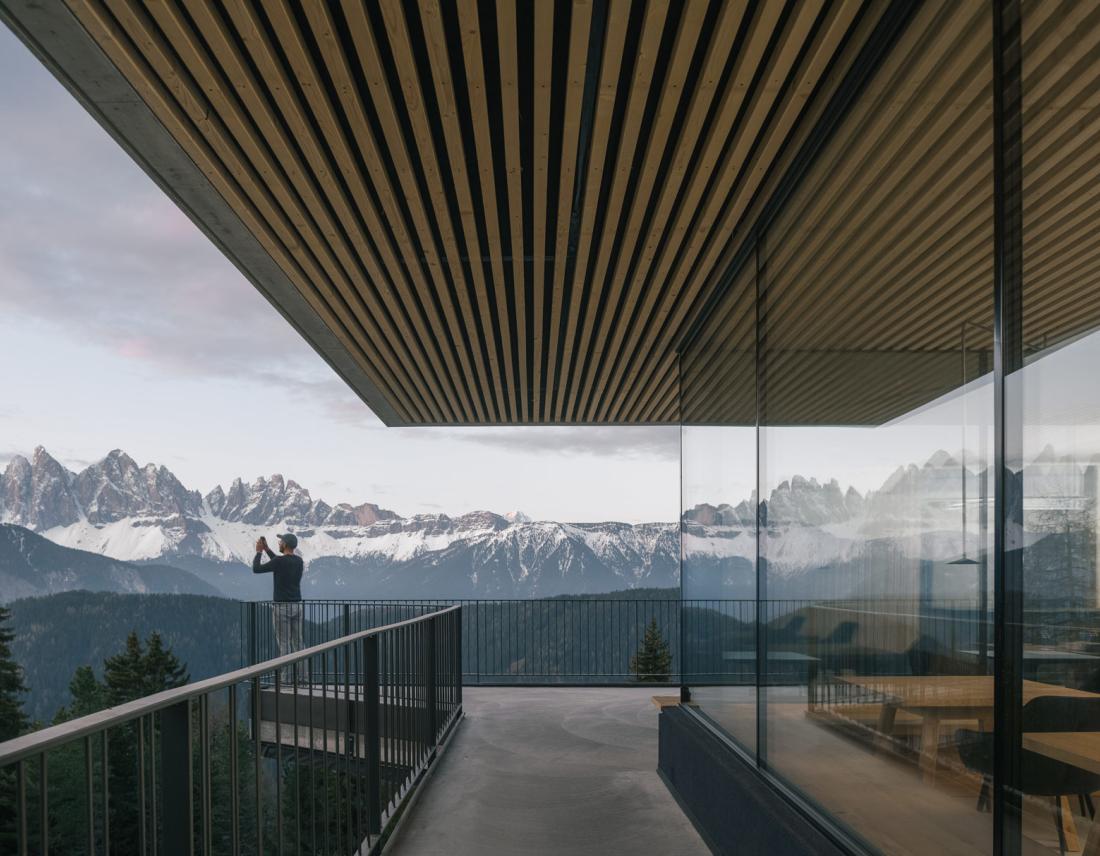 Balkon im Anders Mountain Suites in Brixen: Moderne Terrasse mit Holz und Glas und Blick auf die verschneiten Dolomiten. Ein Mann steht auf dem Balkon und macht ein Foto von der Aussicht.