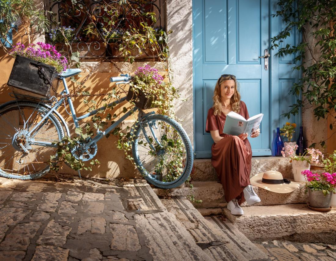 Silvia Trippolt-Maderbacher, die in Istrien auf dem Boden vor einer blauen Türe sitzt mit einem Buch in der Hand. Daneben steht ein blaues Fahrrad