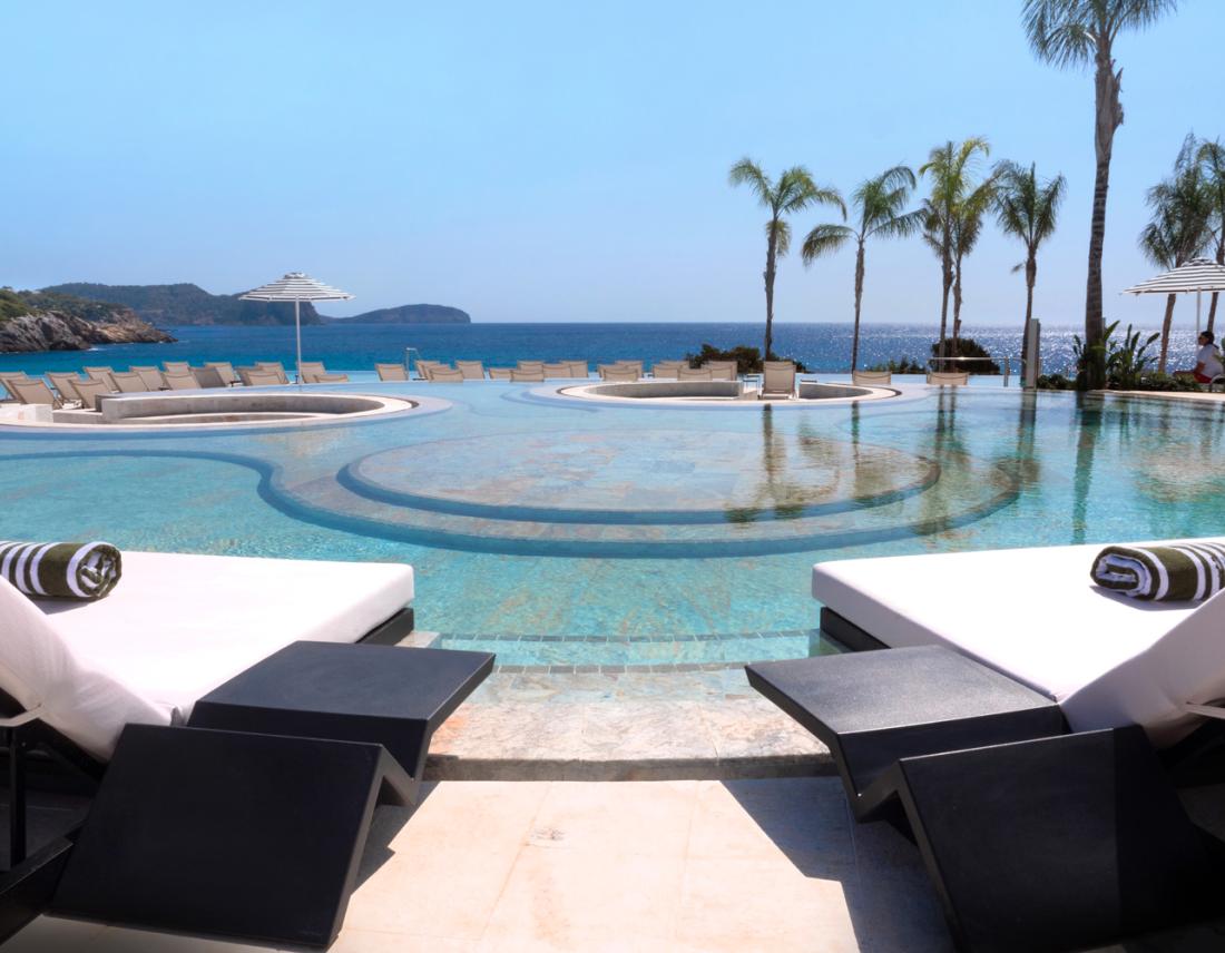 Pool Area mit großem runden Pool und Liegestühlen. Im Hintergrund sind Palmen und das Meer.