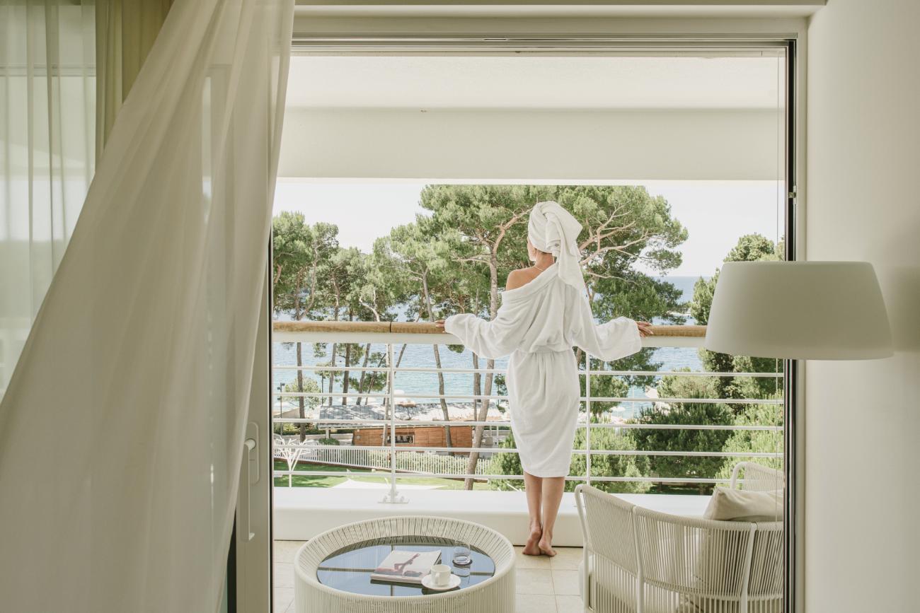 Frau im Bademantel mit Handtuch auf dem Kopf steht auf einem Balkon und blickt auf Bäume und das Meer