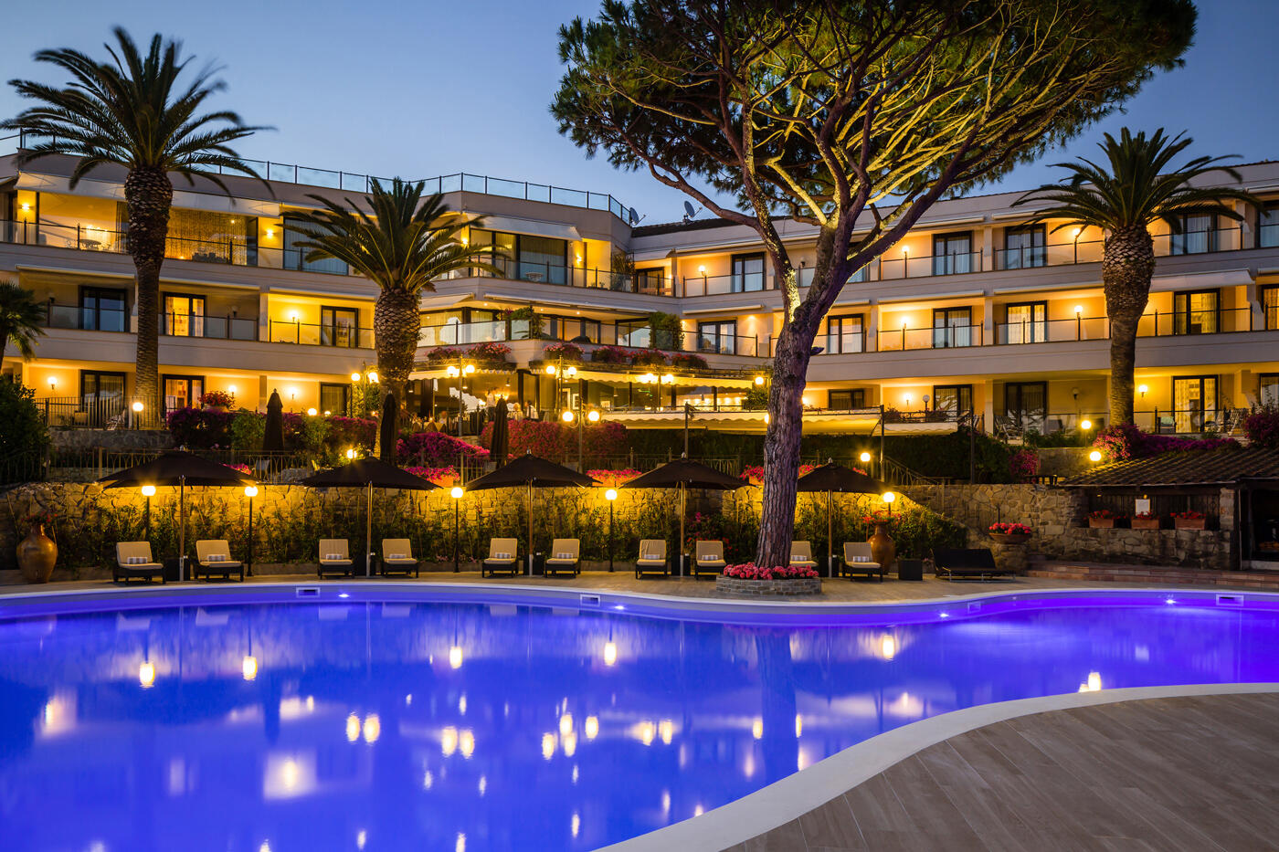 Hotelfassade vom Baglioni Resort Cala Del Porto am Abend mit Pool und Pinienbäume im Vordergrund.