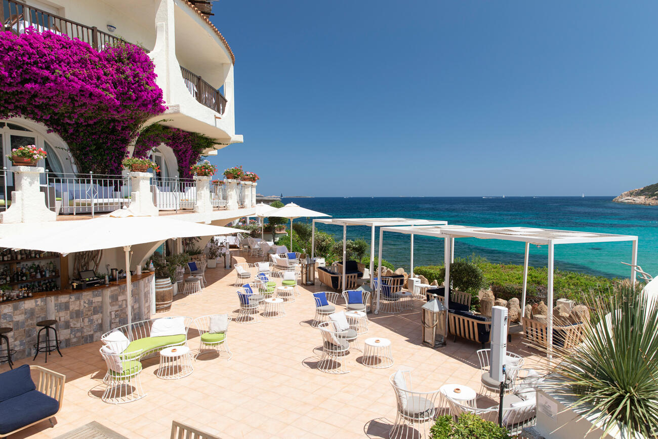 Terrasse mit Tischen, Stühlen und Schirmen. Links im Bild ist eine weiße Hausfassade mit lila Blumen. Rechts sieht man das türkise Meer.