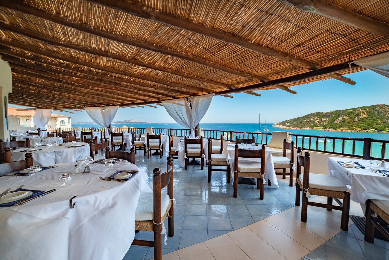 Überdachte Terrasse eines Restazrants mit vielen Tischen und Stühlen. Im Hintergrund sieht man das Meer.