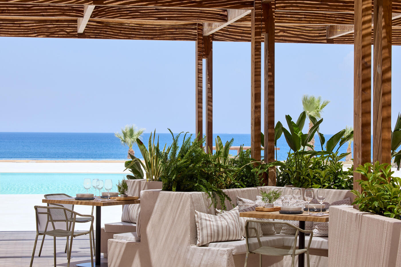 Mediterranes Restaurant mit Sonnendach und vielen Pflanzen direkt am Pool. Im Hintergrund sieht man das Meer.