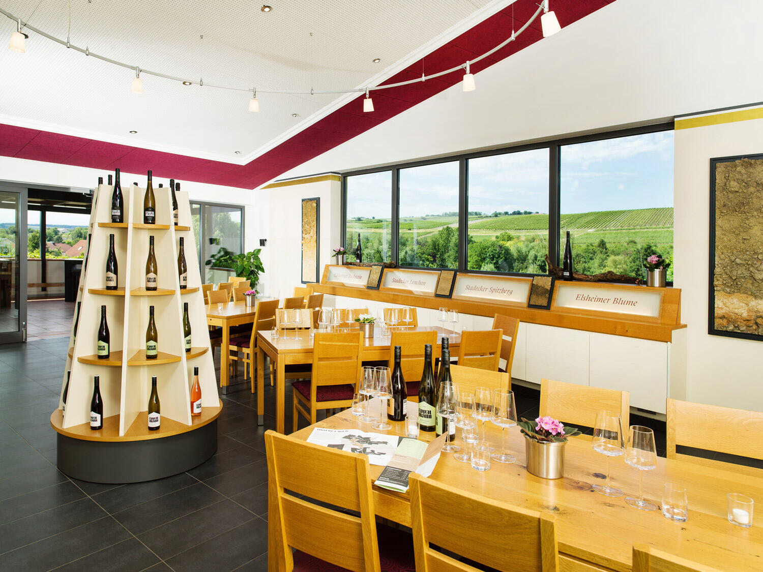 Vinothek in Rheinhessen mit hellen Holzitschen und Stühlen und großen Fenstern mit Weingartenblick. Auf den Tischen stehen Gläser und Weinflaschen.