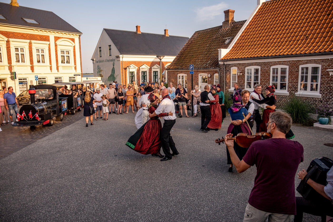 Menschen in tradizionellen Gewändern beim Tanzen auf einen großen Platz. Im Hintergrund stehen bunte Häuser.