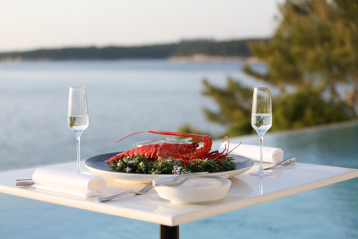 Weißer Tisch mit einem Teller und zwei Weingläser. Im Hintergrund ist ein Pool und das Meer.