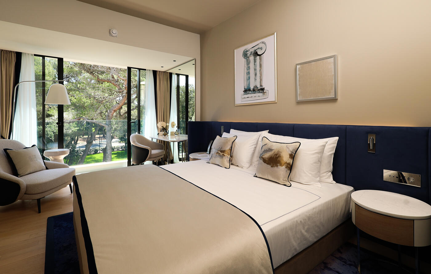 Hotelschlafzimmer mit Doppelbett und beigen Tönen. An der Wand hängen zwei Bilder. Im Hintergrund ist eine offene Terassentüre.