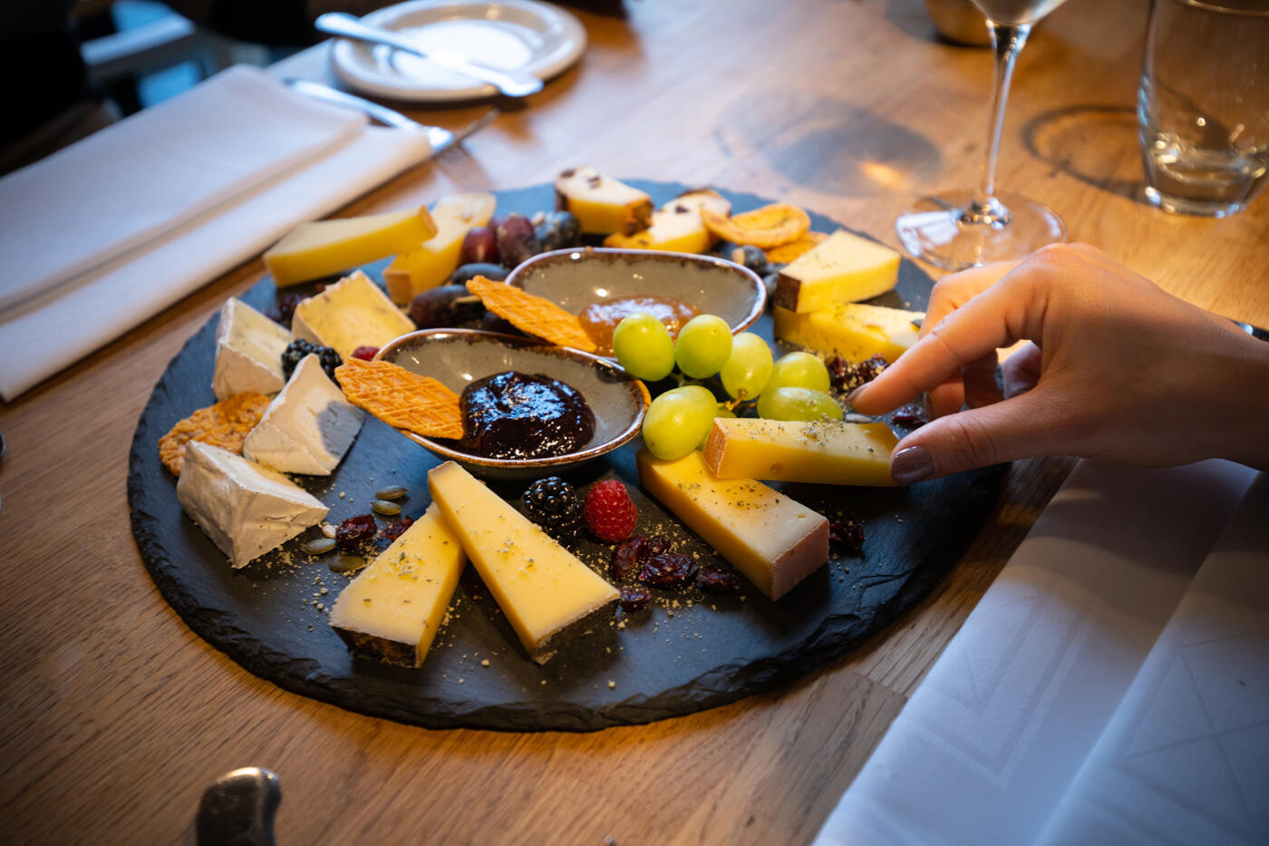 Teller mit Käsevariationen und Trauben. Rechts ist eine Hand, die ein Stück Käse greift.
