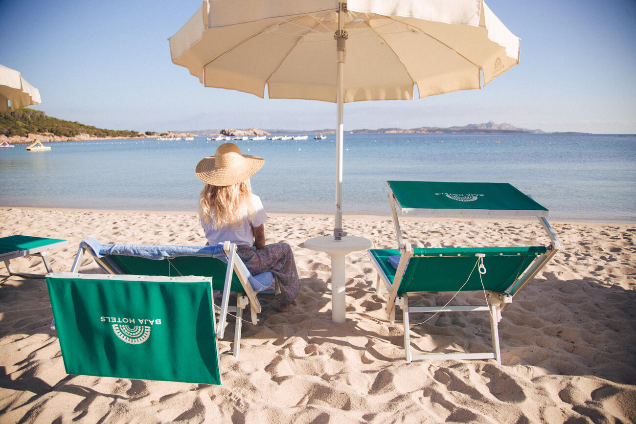 Frau mit Hut, die unter einem weißen Schirm auf einer grünen Liege am Strand sitzt. Neben ihr steht eine weitere grüne Sonnenliege. Im Hintergrund ist das Meer.