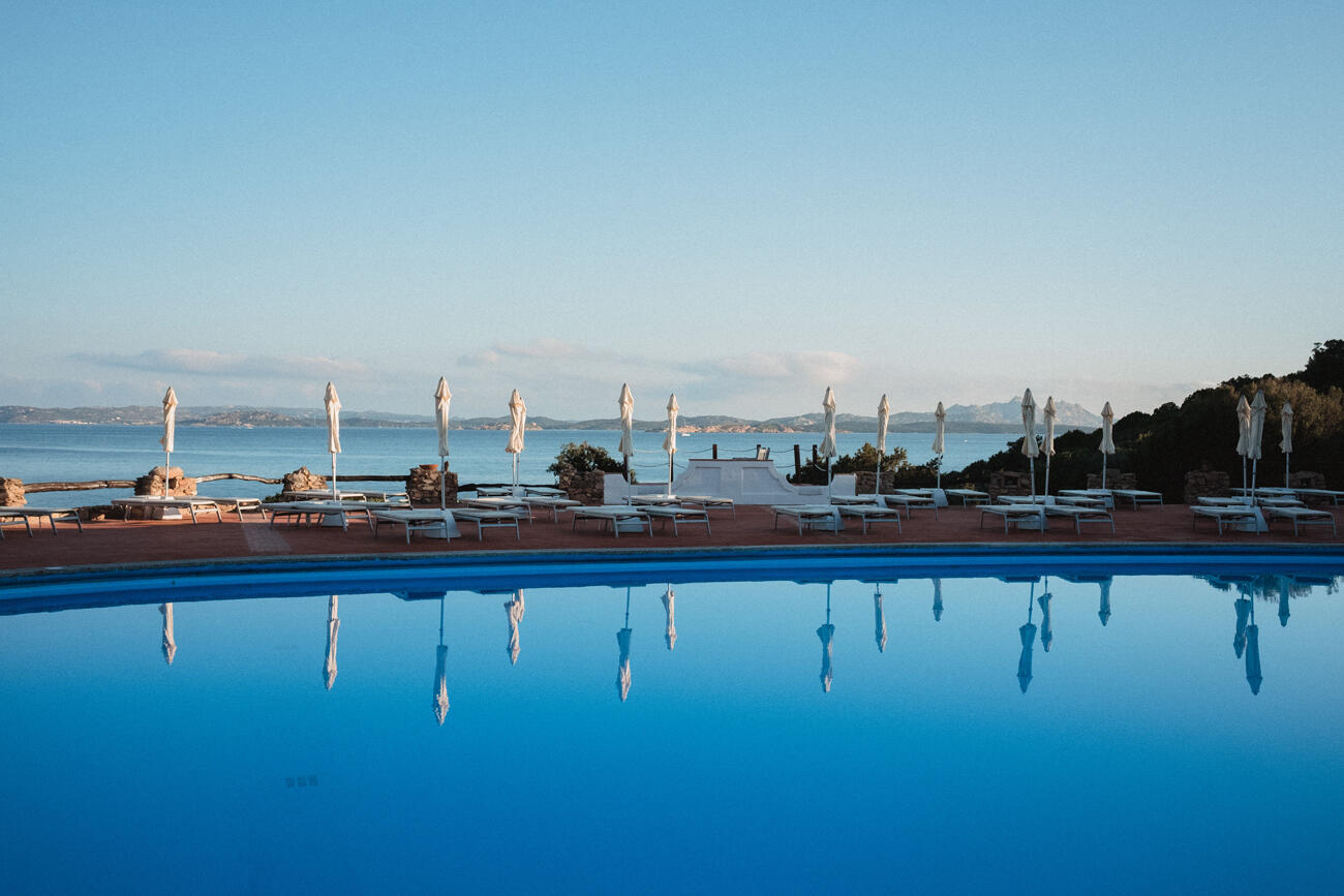 Pool mit Liegestühlen und geschlossenen Schirmen in der Abendstimmung. Im Hintergrund sieht man das Meer.