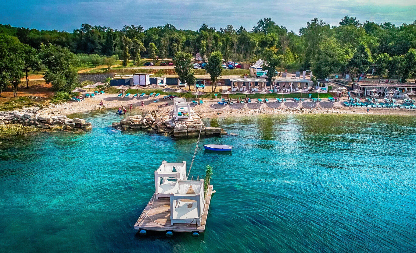 Blick auf Strand Club in Istrien. Vorne sieht man das Meer, im Hintergrund Sonnenliegen und Schirme.