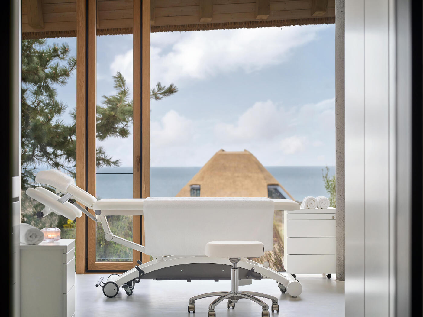 Treatmentraum im Lanzerhof: weiße Behandlungsliege in einem hellen Raum mit großen Glasfenstern und Meerblick