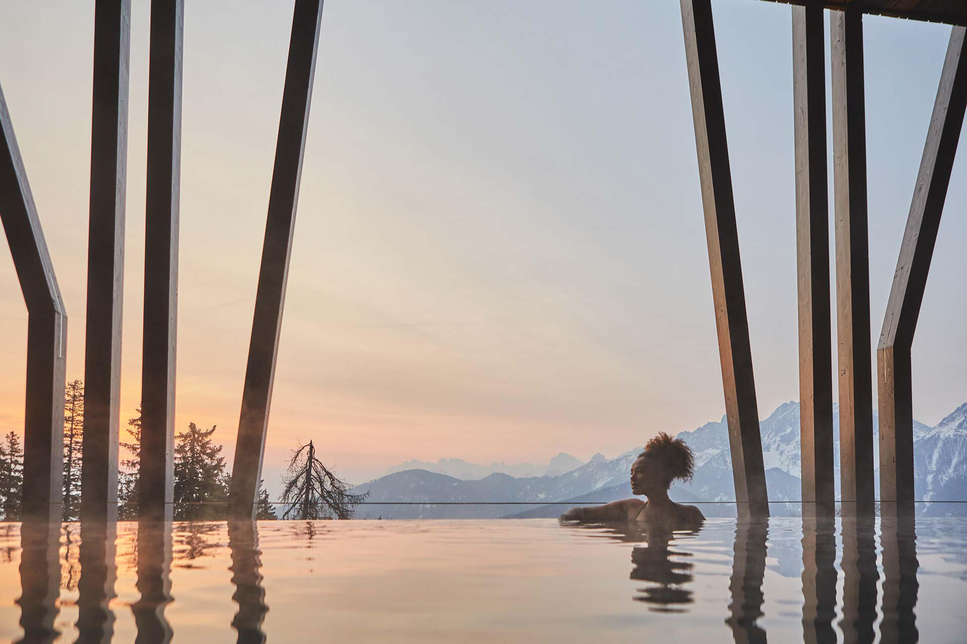 Frau in einem Infinity Pool während des Sonnenuntergangs. Im Hintergrund sind Berge.