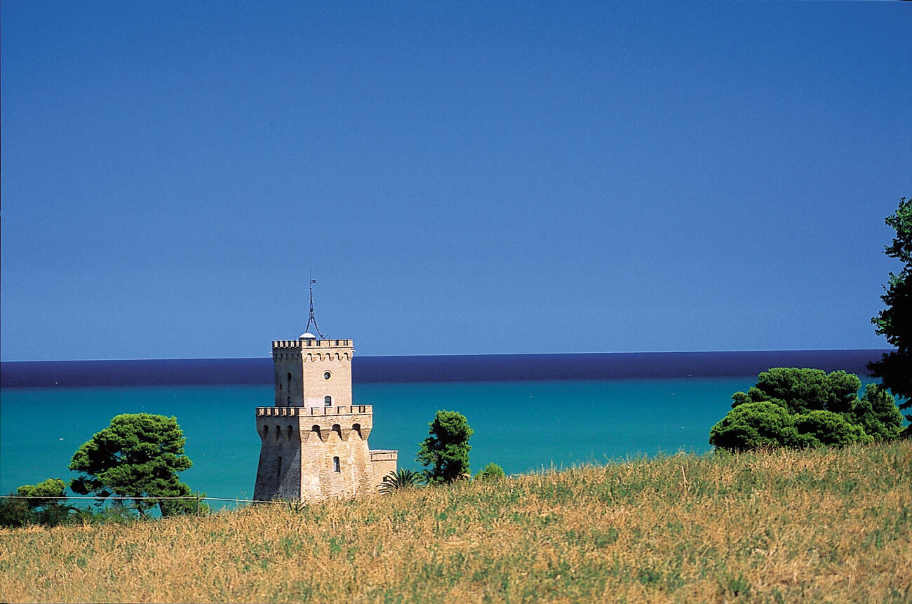 Ein altertümlicher Turm der vor dem türkisfarbenen Meer steht. Im Vordergund ist eine bunte Wiese.