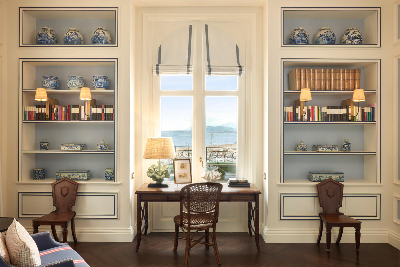 Blick auf eine Wand mit zwei Einbauregalen jeweils auf einer Seite. In der Mitte sieht man ein Fenster mit Meerblick. Davor steht ein kleiner Tisch mit Stuhl und Lampe.