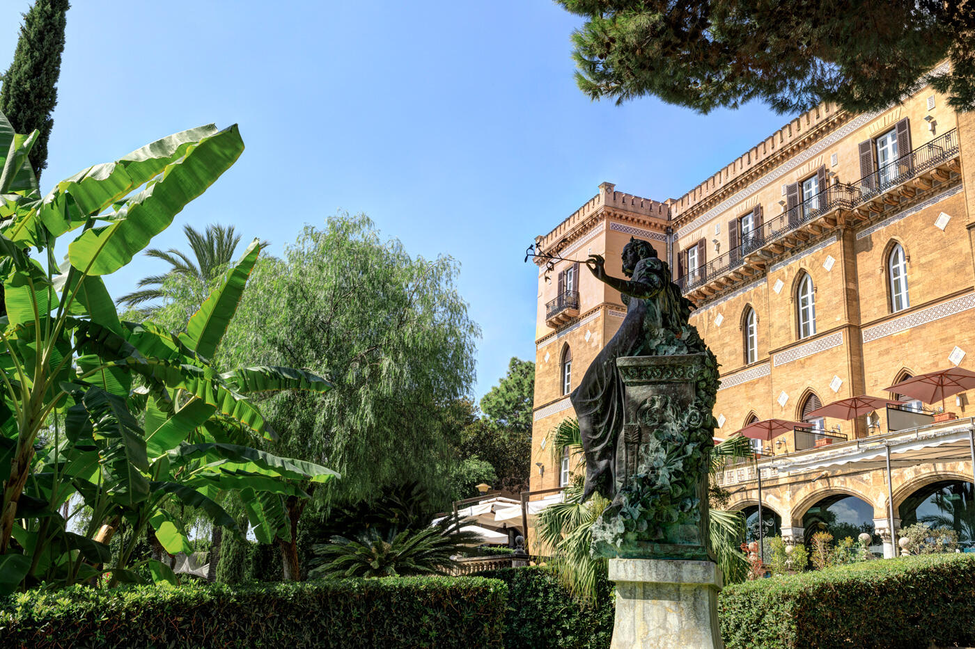 Villa Igiea im Jugendstil mit hellbrauner Fassade. Drumherum wachsen grüne Pflanzen. im Vordergrund sieht man eine Statue.