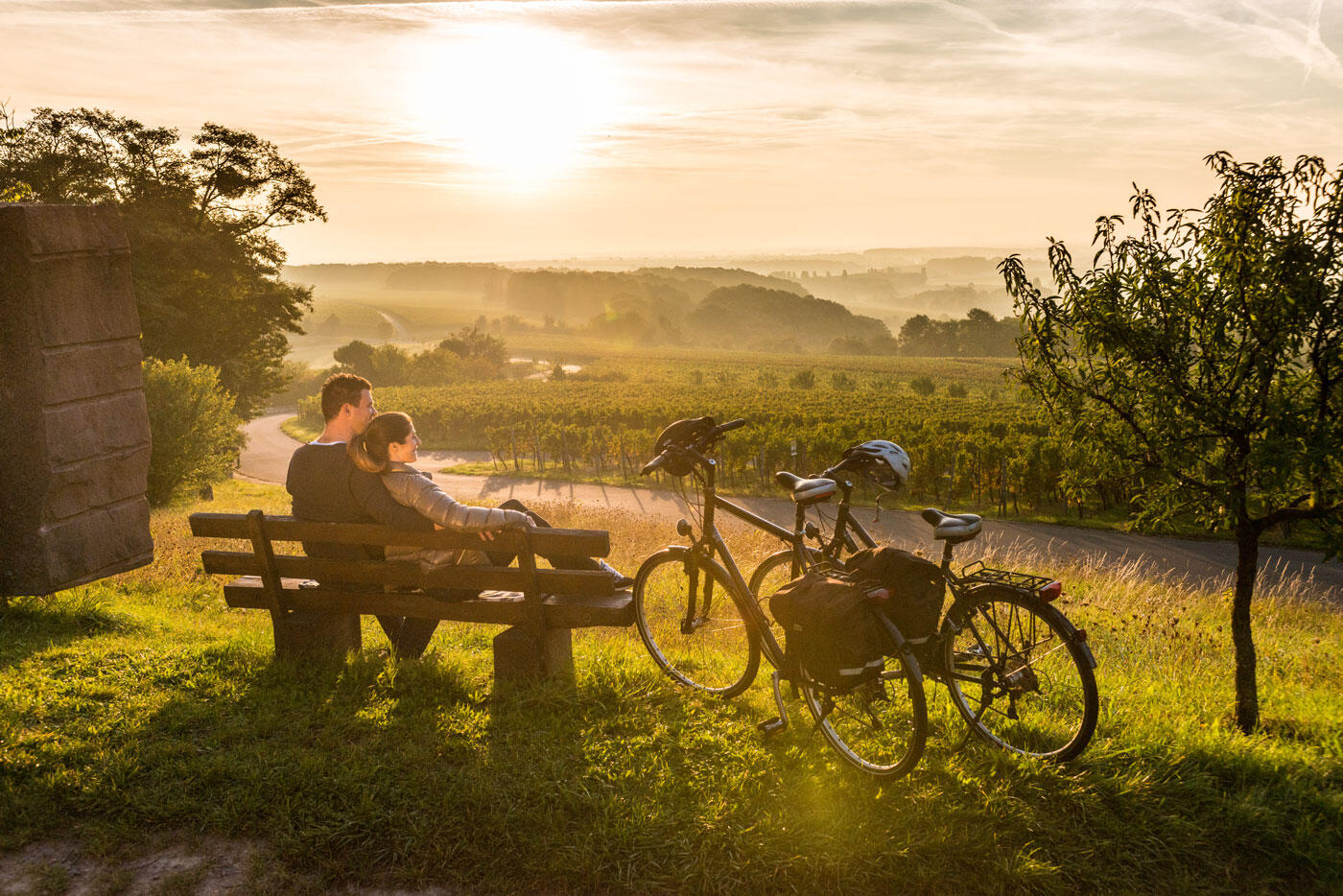 Pärchen, dass auf einer Bank sitzt und den Sonnenuntergang über den Weinbergen beobachtet. Am Rand stehen zwei Fahrräder.