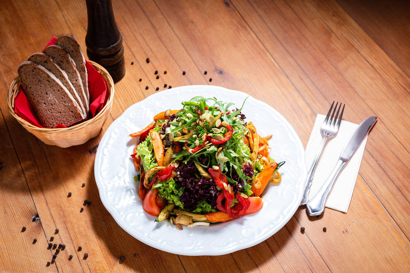 Bunter Salat auf einem weißen Teller. Daneben steht ein Korb mit mehreren Brotscheiben. Im Hintergrund sieht man einen Holztisch