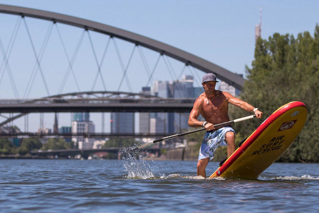 Mann auf einem Stand Up Paddle-Board im Wasser. Im Hintergrund ist eine Brücke und eine Skyline.
