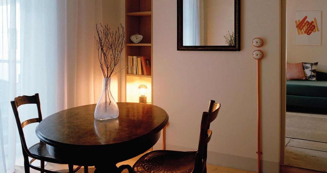 Blick auf runden Holztisch mit Blumenvase und zwei Stühlen und gemütlichem Licht im Steinach Townhouse Meran