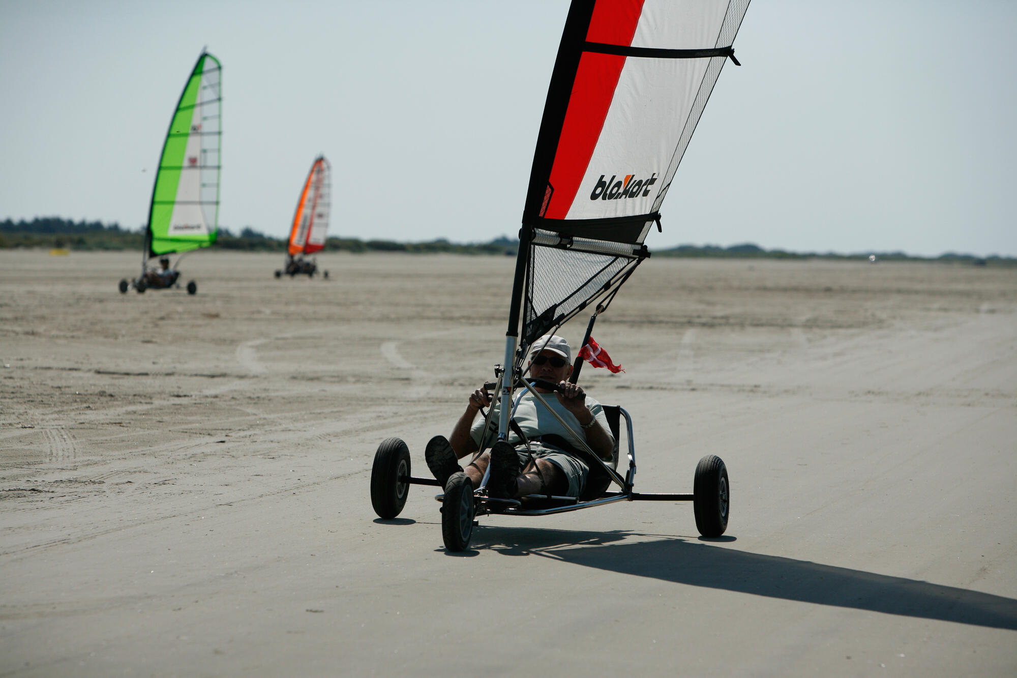 Mann in einem Kitebuggy mit Segel, der über einen leeren Strand fährt. Im Hintergrund sind zwei weitere Kitebuggys