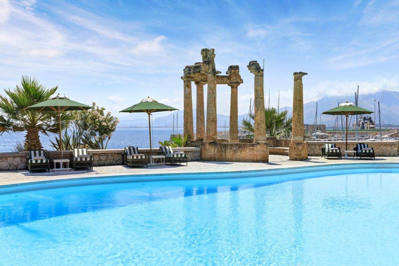 Pool mit Liegestühlen und Sonnenschirmen und römischen Säulen. Im Hintergrund ist der Golf von Palermo
