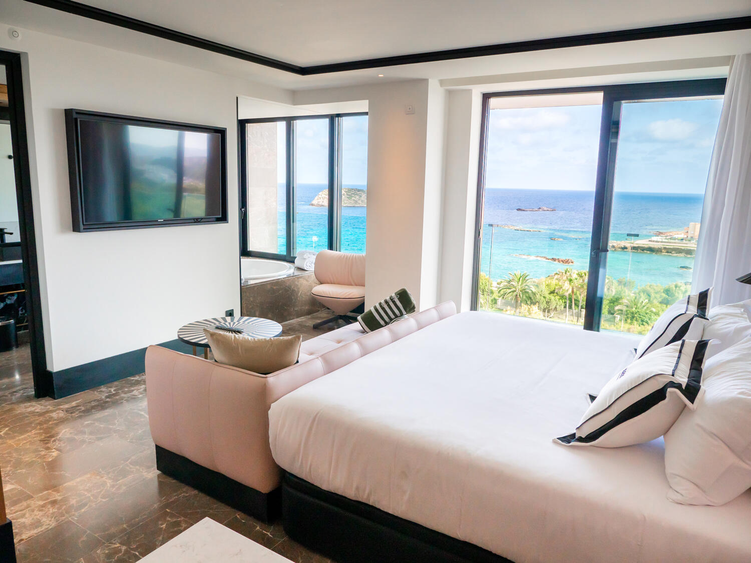 Hotelzimmer im Bless Hotel Ibiza mit Doppelbett und rosaner Couch und großen Glastüren, die Blick auf das Meer geben.