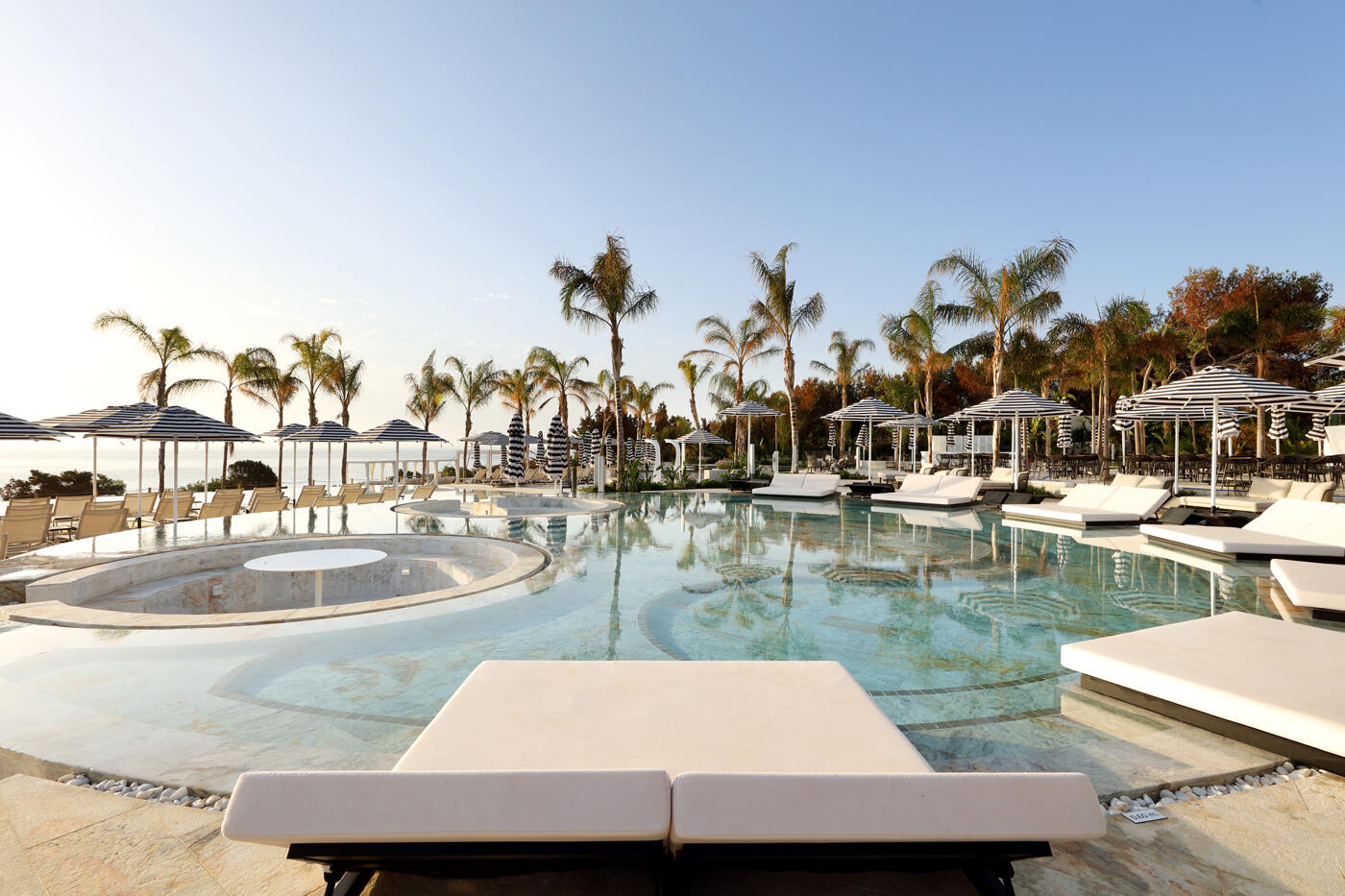 Runder Pool mit weißen Liegestühlen drumherum. Im Hintergrund stehen Palmen und man sieht das Meer.