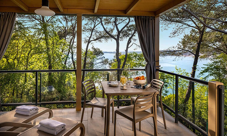 Überdachter Balkon mit Liegestühlen und einem Tisch mit Stühlen. Im Hintergrund sind Bäume und das Meer