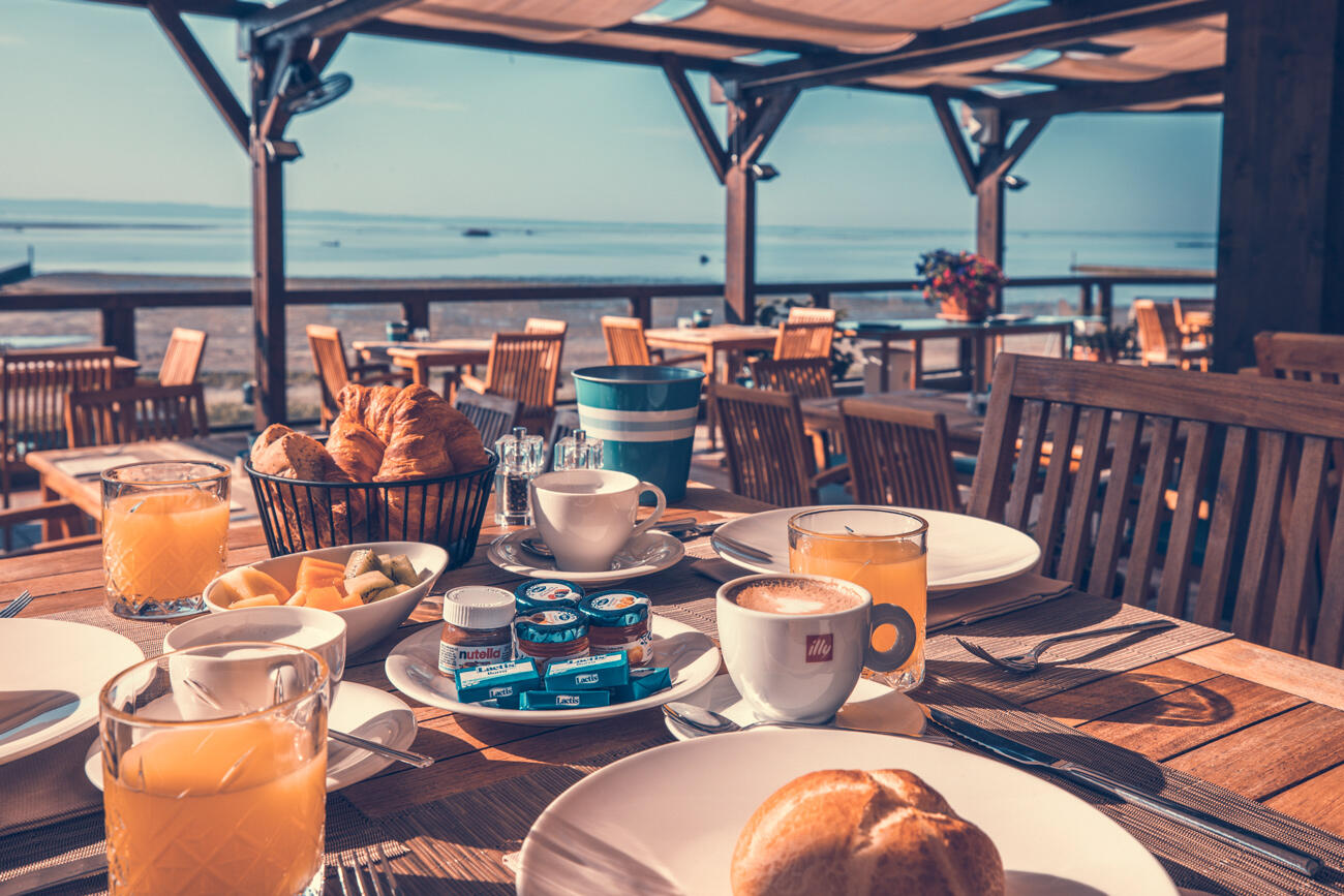 Große Restaurant Terrasse direkt am Strand. Im Hintergrund sieht man das Meer, im Vordergrund einen gedeckten Holztisch mit Kaffee, Orangensaft, Brot und Nutella