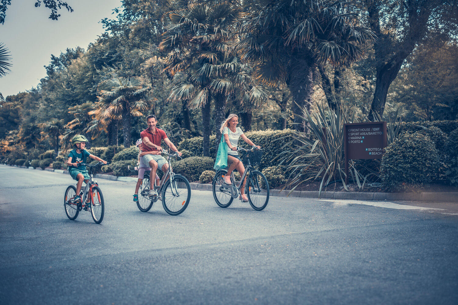 Eine Frau, ein Mann und ein Kind, die auf einer Terrstraße mit dem Fahrrad fahren. Im Hintergrund wachsen Palmen, Bäume und Büsche
