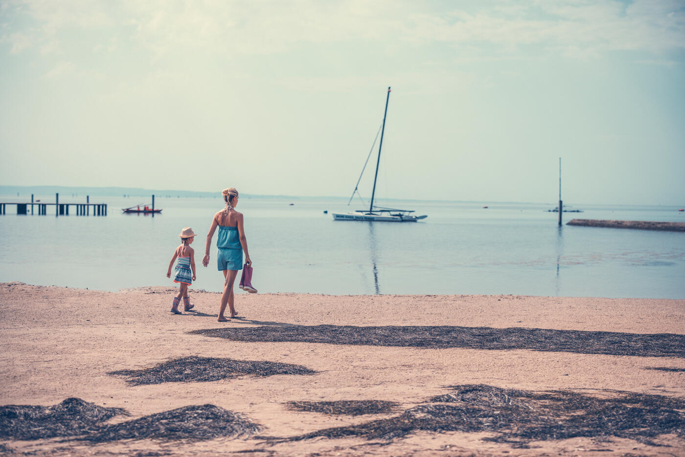 Frau mit Kind, die an einem Strand in Grado spazieren. Im Hintergrund sieht man ein Segelboot.